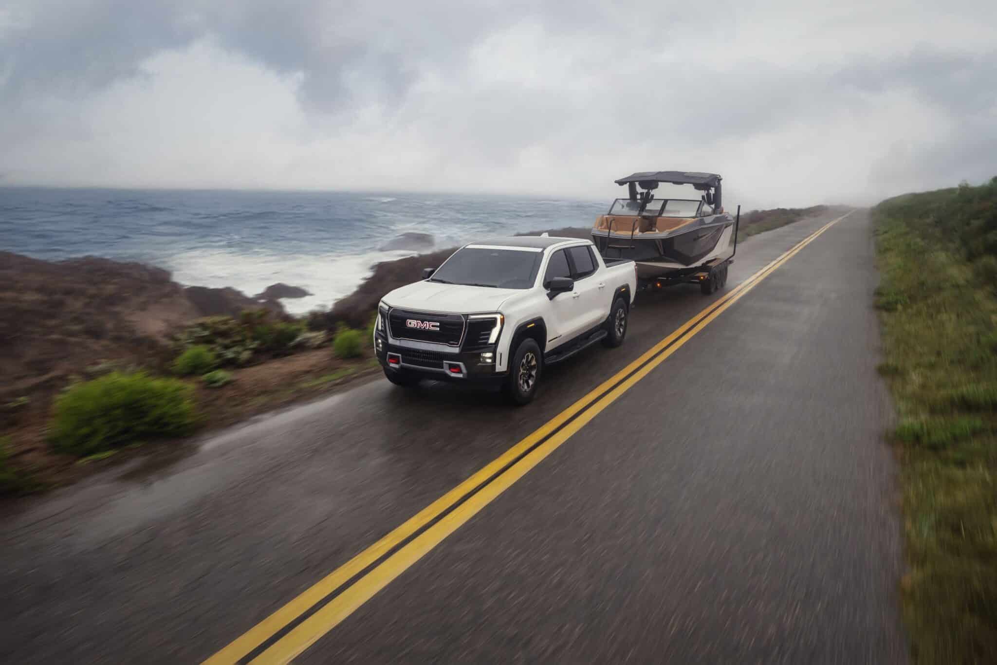 Front 3/4 view of the 2026 GMC Sierra EV AT4 in Summit White towing a boat on a seaside road.